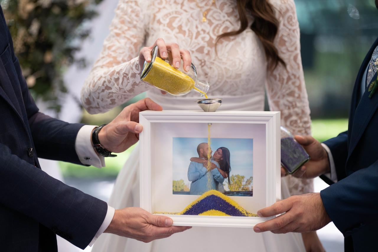 Dos hombres sujetan un marco mientras una chica vestida de novia vierte en su interior arena en color amarillo