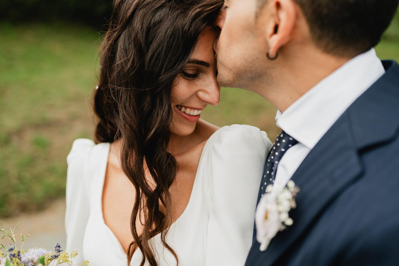 Chica morena con vestido blanco y su melena morena suelta sonríe feliz mientras un chico con traje le da un cariñoso beso en la frente