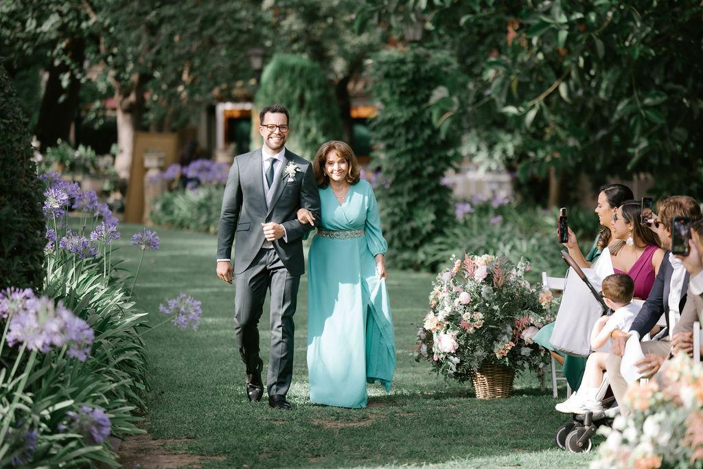 Madrinas de boda: novio con traje y madrina con un vestido largo en color aguamarina caminan por el pasillo de su ceremonia de boda civil al aire libr