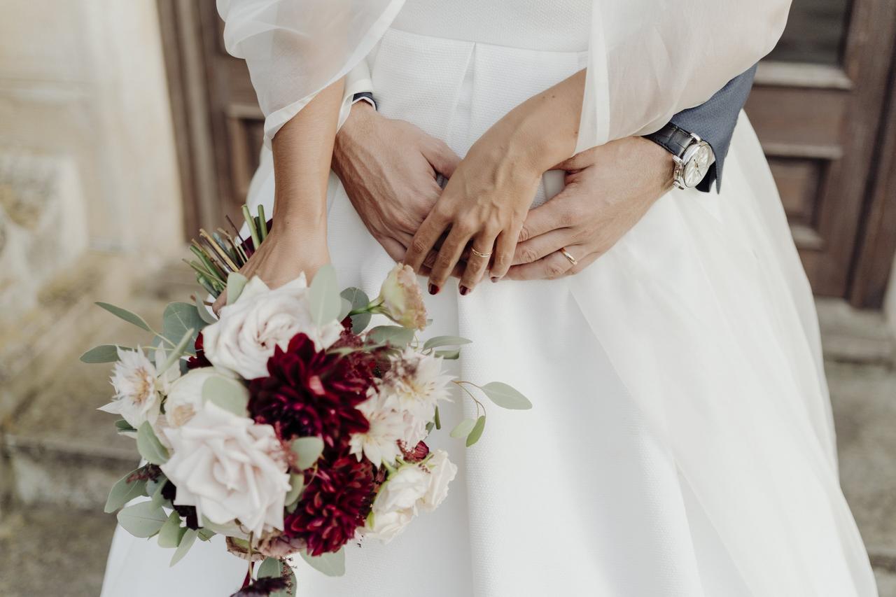 Diferencia entre pareja de hecho y matrimonio: chica con un vestido blanco y un ramo de novia en una mano coge con la otra las manos de un hombre, que la abraza por detrás