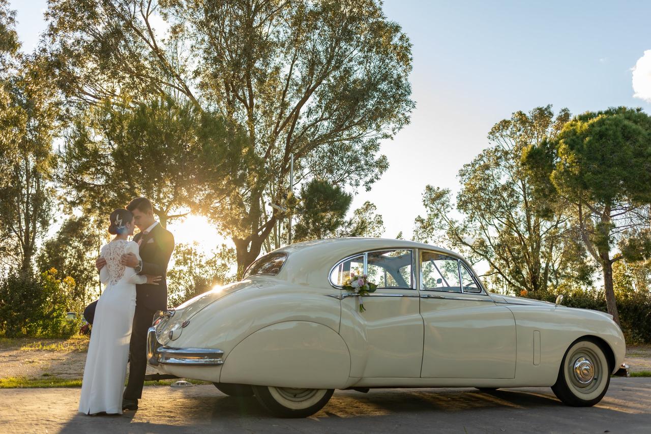 Pareja vestida de novios se abraza apoyada en un precioso coche clásico de color claro decorado con flores