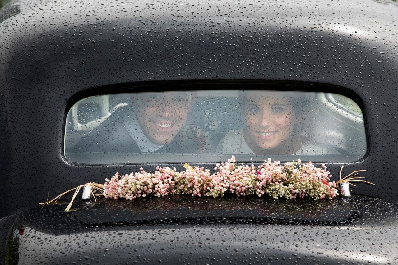 Boda con lluvia: chico y chica en el interior de un coche clásico mirando por el cristal trasero salpicado de lluvia