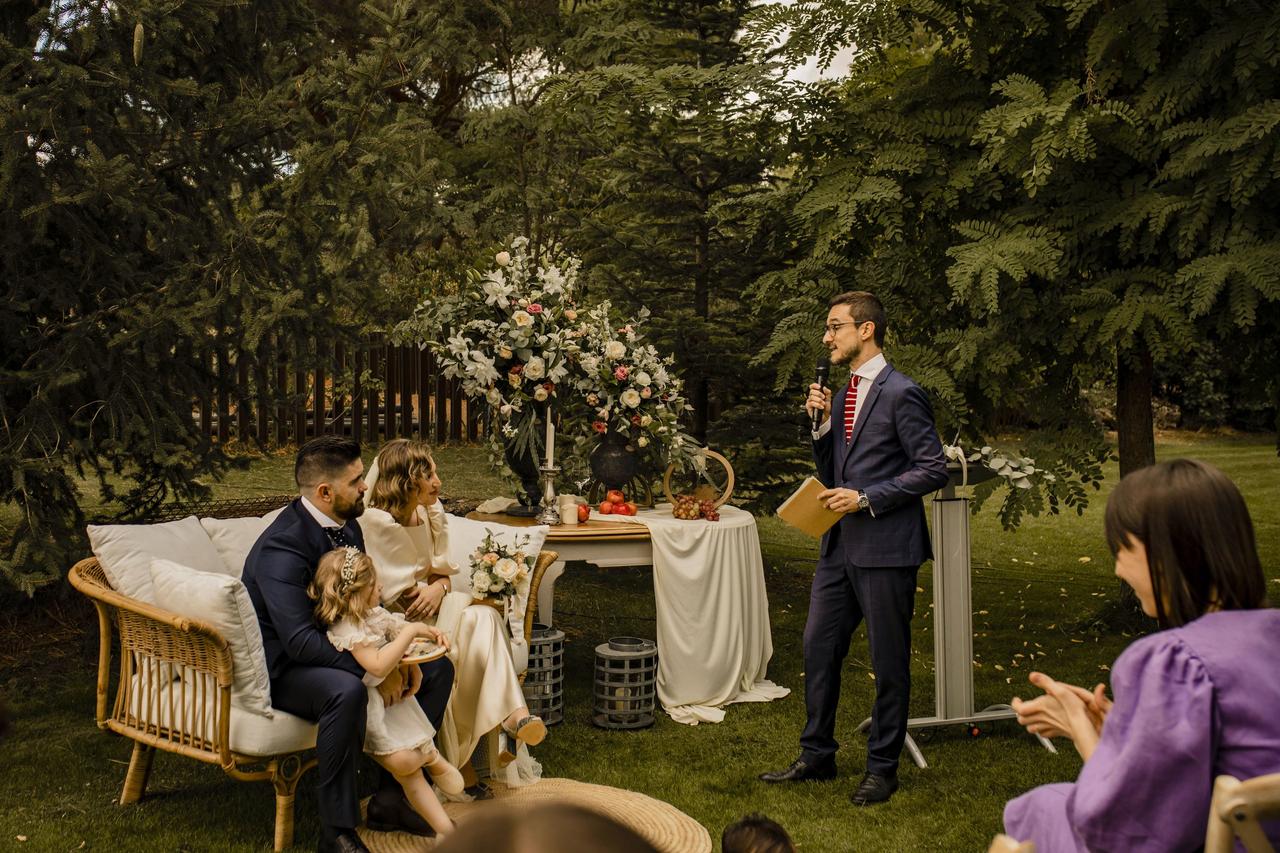 Ceremonia de boda civil en un jardín con mucho verde con los contrayentes sentados en un banco con una niña y el oficiante hablando con el micrófono en la mano