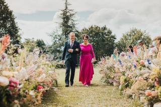 Madrinas de boda: novio con traje clásico y madre con vestido fucsia largo y clutch en la mano caminan por el pasillo de su boda civil al aire libre