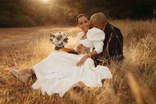 Chico con traje y chica con vestido de novia sentados en un campo mientras él la abraza a ella por detrás