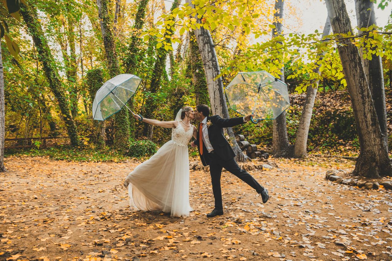 Boda con lluvia: chica vestida de novia y chico con traje oscuro en una divertida foto en medio de la naturaleza y protegiéndose de la lluvia con paraguas transparente