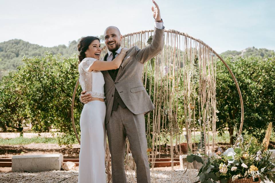 Chico con traje beige y chica vestida de novia se abrazan muy sonrientes junto al altar de su boda civil al aire libre