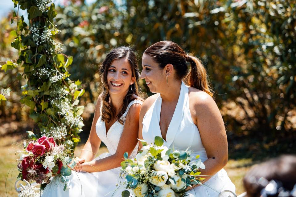 Dos chicas sonrientes, vestidas de novia, sentadas en el altar con un ramo de flores cada una en las manos