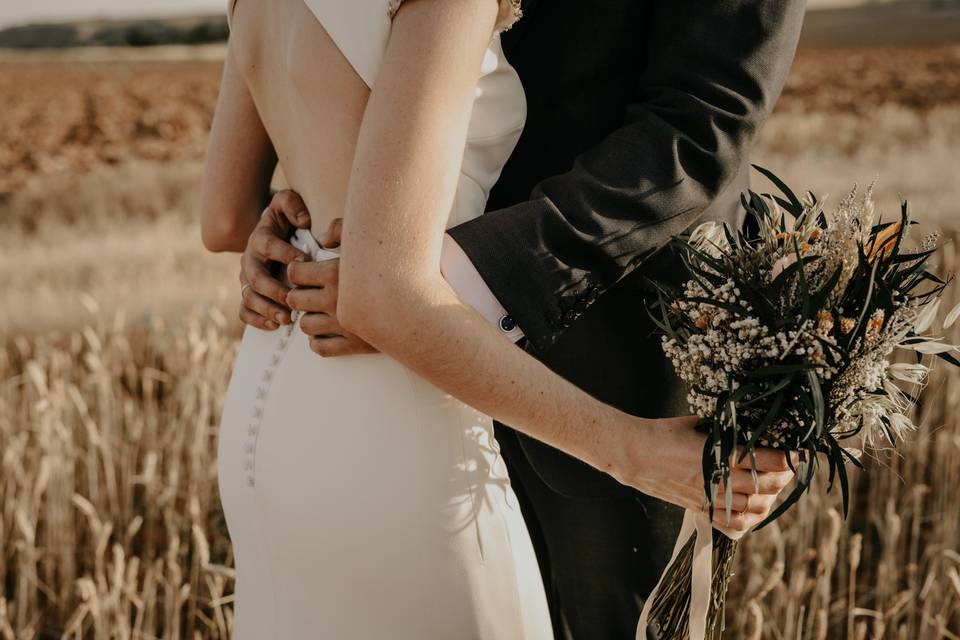 Chica con un vestido blanco y un ramo de flores en una de sus manos y chico con traje se abrazan en un campo de trigo