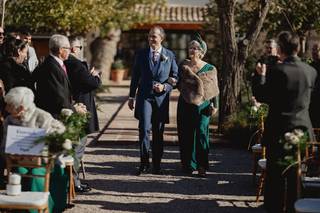 Madrinas de boda: novio con traje y madrina con mono verde y estola de piel caminan por el pasillo de su ceremonia civil al aire libre