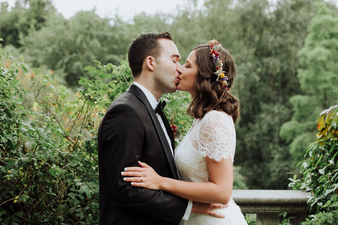 Chica vestida de novia con media melena y una corona de flores en la cabeza se da un beso con un chico con traje clásico