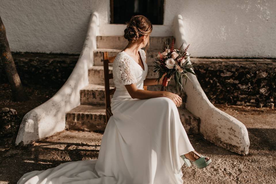 Novia sentada de lado en una silla de madera en el exterior mientras sujeta con una de sus manos su ramo de novia