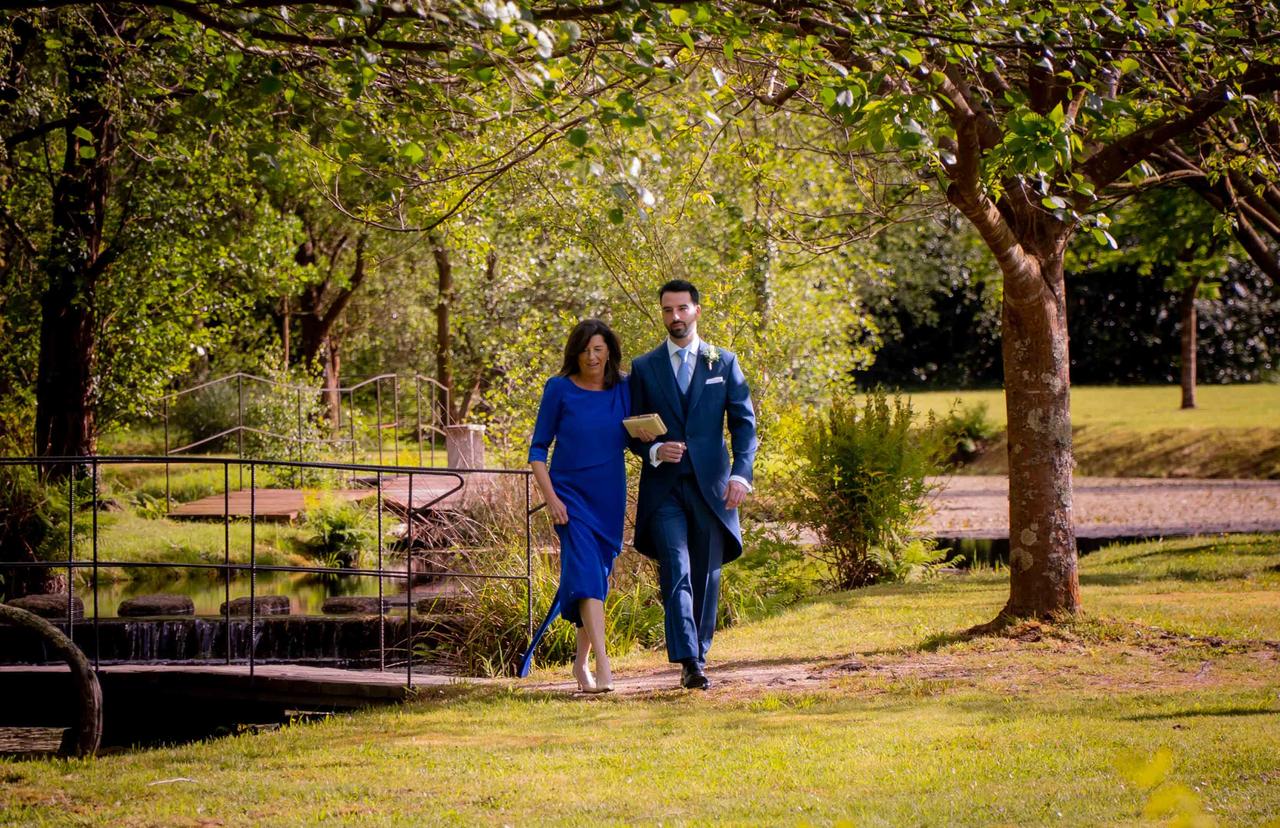 Madrinas de boda: novio con traje y madrina con vestido azul de largo midi caminan cogidos del brazo por un ambiente al aire libre