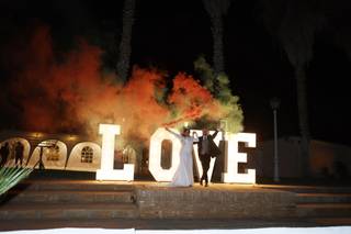 Pareja de recién casados con bengalas de humo de colores en las manos delante de unas letras gigantes iluminadas para boda con la palabra Love