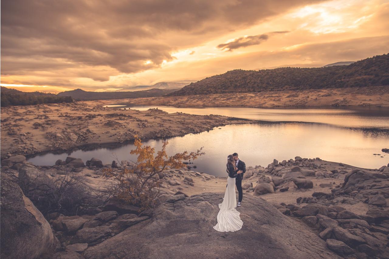 Chico y chica con vestido de novia y chupa de cuero se abrazan junto a un embalse durante la puesta de sol