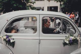 Wolkswagen escarabajo con una novia sentada en el asiento de atrás muy sonriente y con flores colgando de las manijas de las puertas