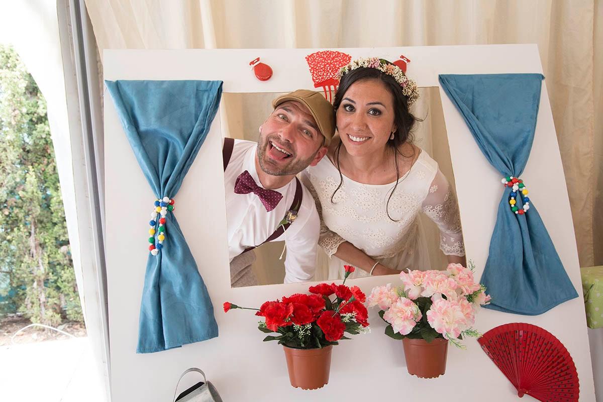 Pareja de recién casados posa divertida en un photocall con forma de ventana con visillos azules y plantas colgando