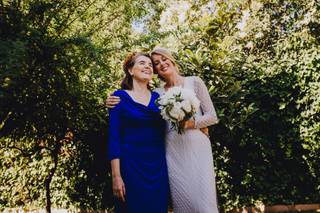 Vestidos para la madre de la novia: chica vestida de blanco y mujer más mayor vestida de azul se abrazan sonrientes al aire libre