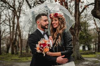 Chica con ramo de flores en una mano y corona de flores sobre su melena suelta posa muy sonriente al lado de un chico con traje que sujeta un paraguas