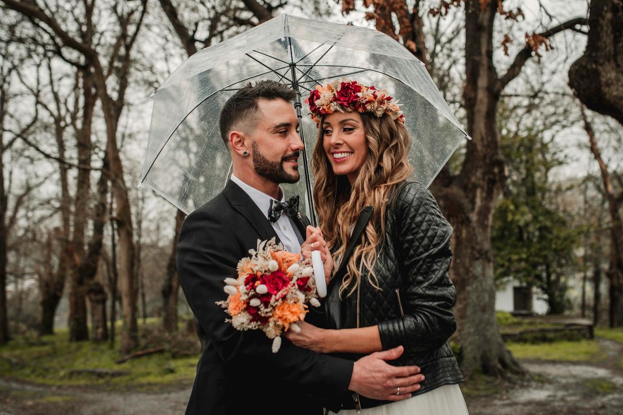 Chica con ramo de flores en una mano y corona de flores sobre su melena suelta posa muy sonriente al lado de un chico con traje que sujeta un paraguas