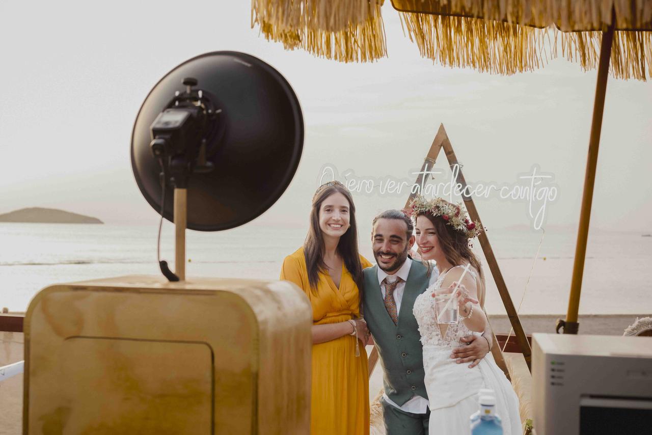 Novia acompañada de un chico y una chica se hace una divertida foto delante de un photocall de boda con el mar al fondo