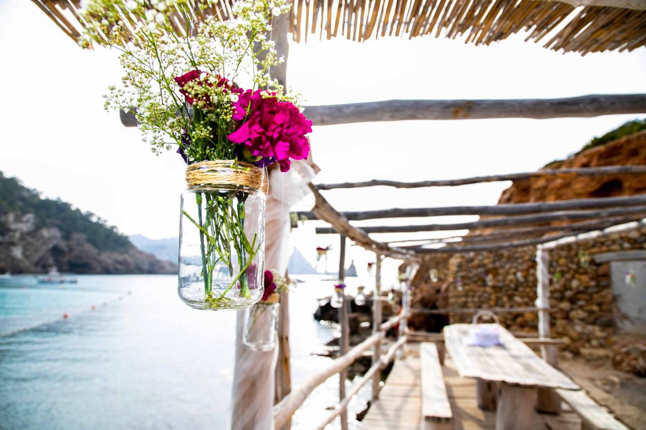 Espacio junto al mar con una larga mesa de madera y bancos del mismo material, con un tarro de cristal con flores en primer plano