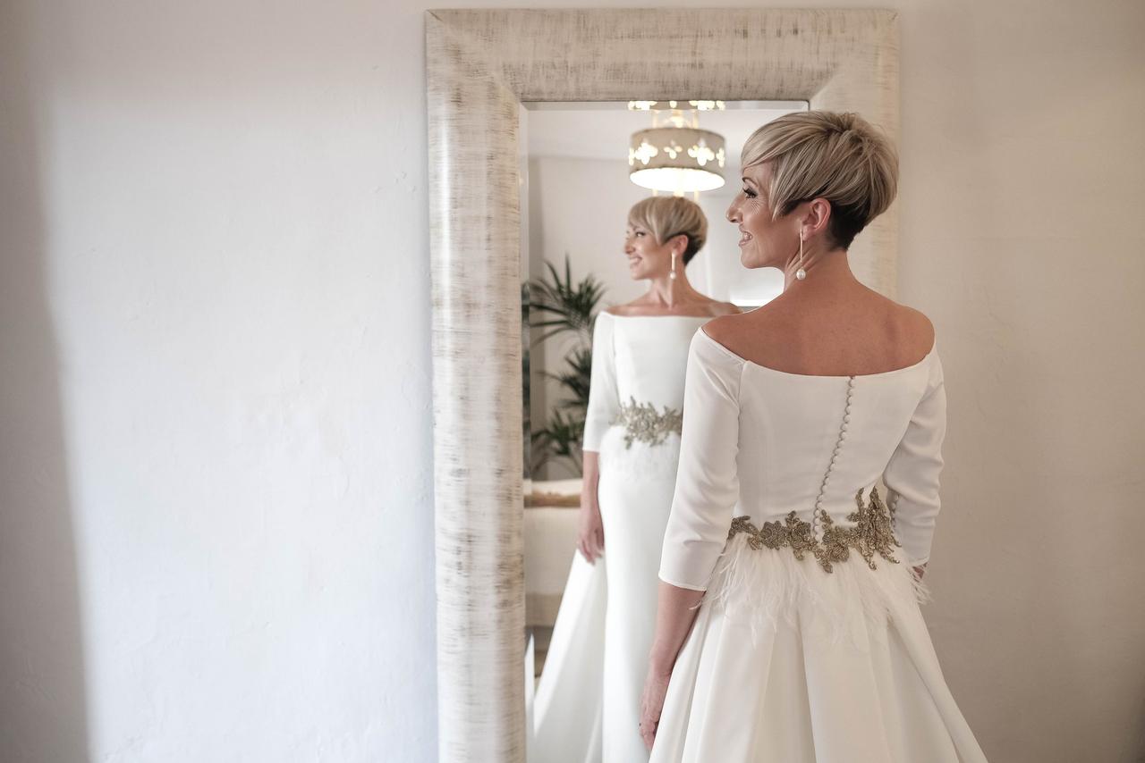 Chica con un vestido de novia con plumas y cinturón de pedrería se refleja en un espejo, mostrando su peinado de novia con pelo corto