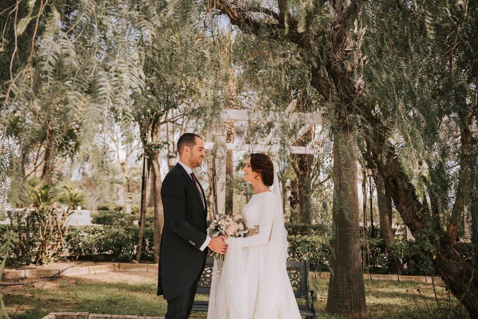 Chico con traje y chica vestida de novia se cogen las manos mientras se miran fijamente a los ojos en un precioso jardín con mucha vegetación