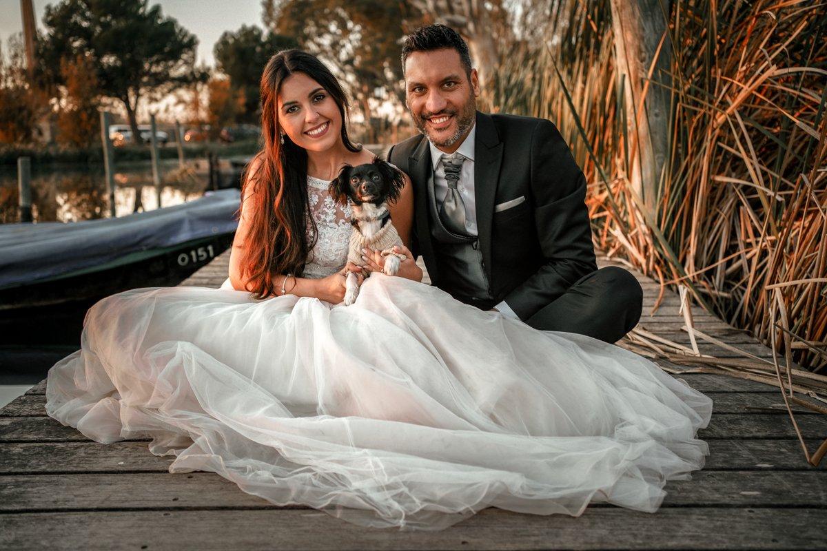 Chica con un vestido de novia y chico con un traje oscuro sentados en un suelo de madera con un perrito