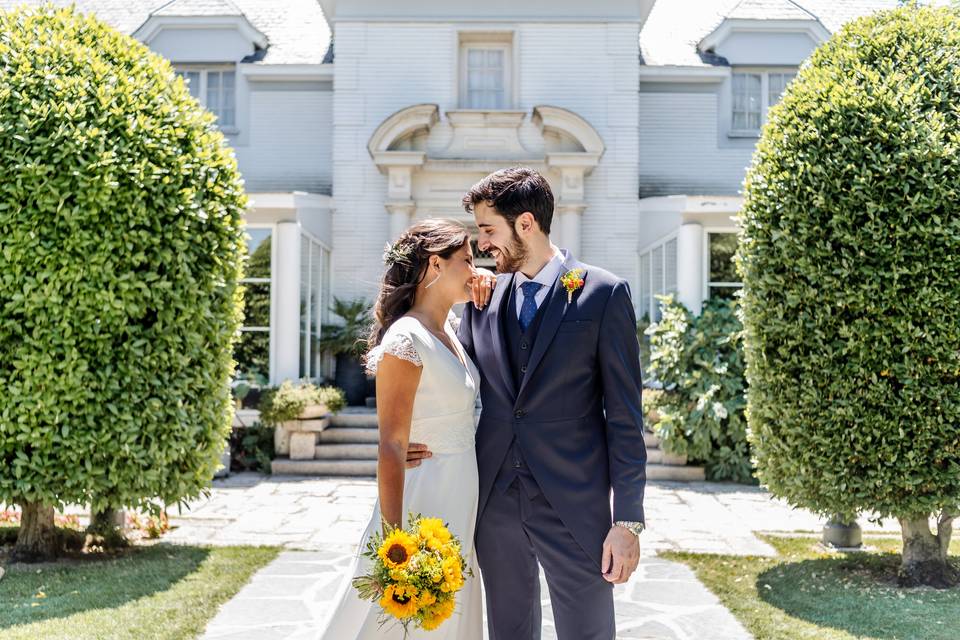 Chico con traje clásico y chica con vestido de novia y ramo de girasoles en la mano posan muy juntos y sonrientes en un precioso jardín