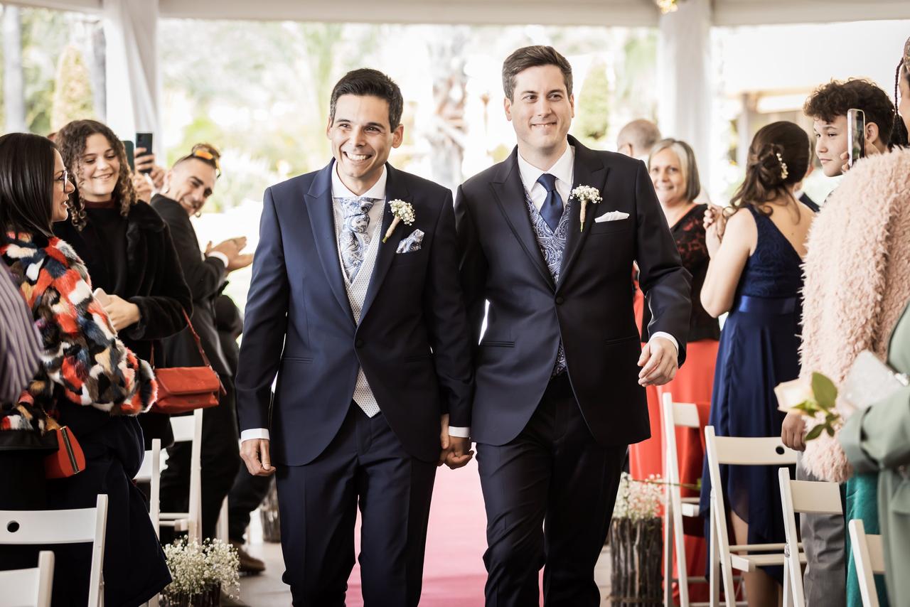 Dos chicos con traje y muy sonrientes caminan cogidos de la mano por el pasillo de su ceremonia de boda civil rodeados de familiares y amigos