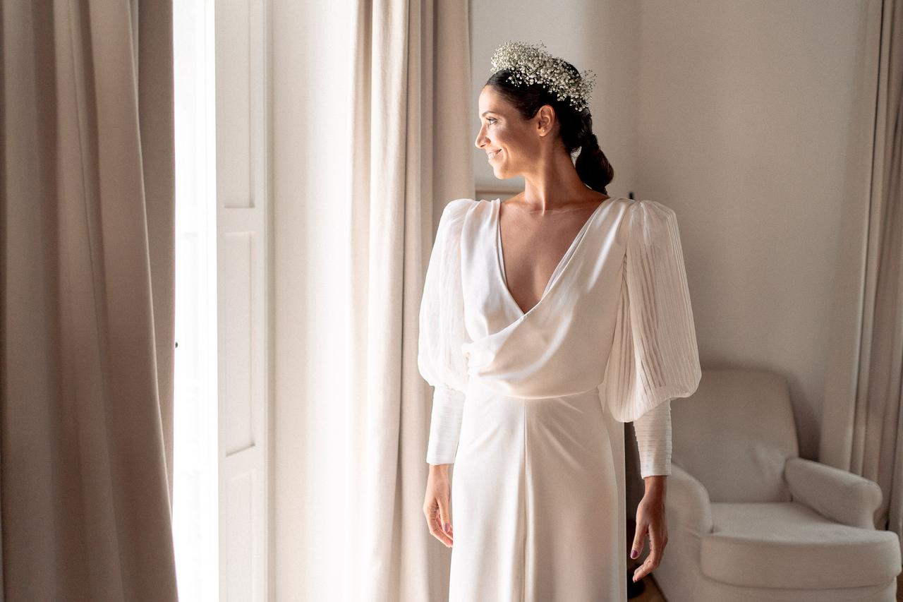 Novia con un precioso vestido con escote en V y manga larga mira sonriente por la ventana de una habitación