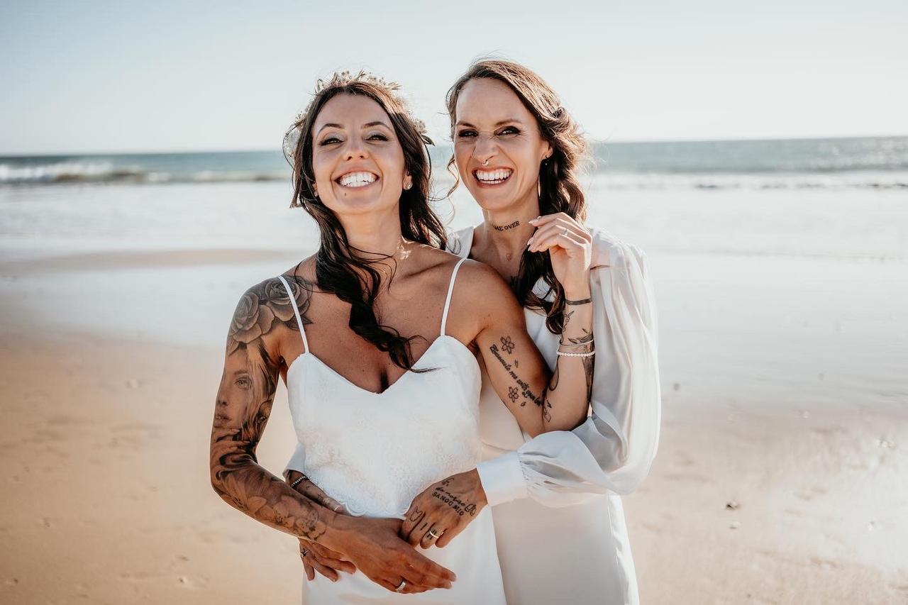Dos chicas muy sonrientes vestidas de novia posan felices en una playa