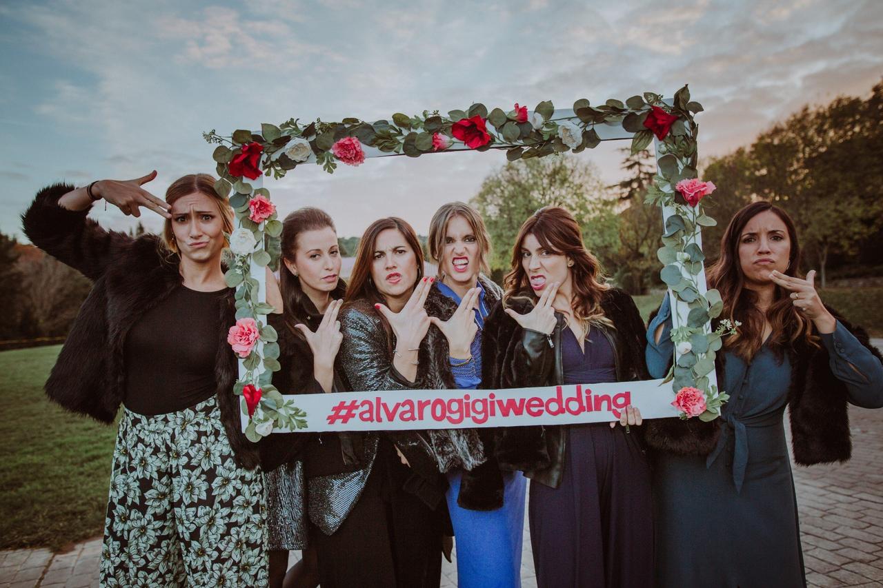 Seis chicas con cara de enfadadas sujetan un marco con flores que hace las veces de photocall de boda