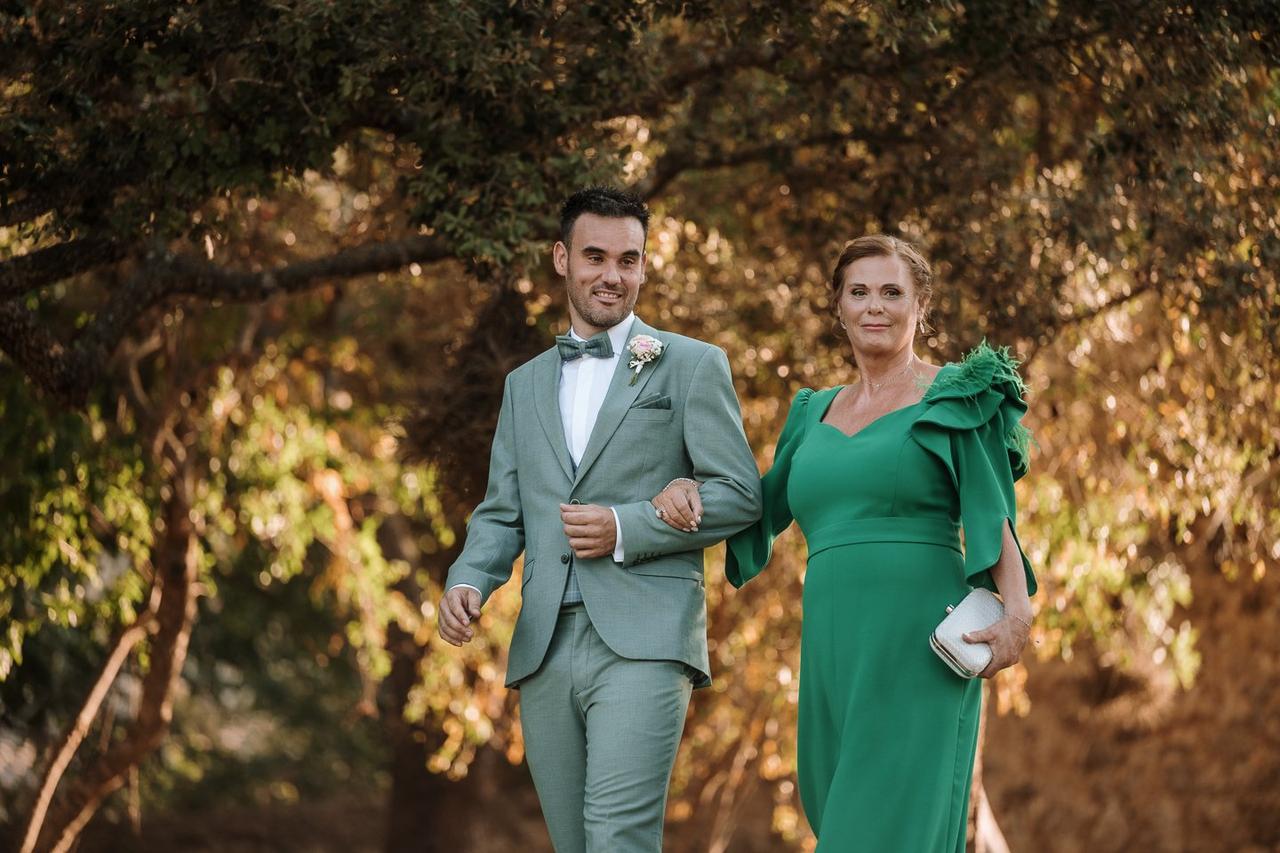 Madrinas de boda: novio con traje verdoso y madrina con vestido verde caminan cogidos del brazo por un exterior