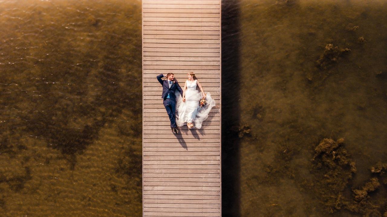 Chico con traje y chica vestida de novia estirados sobre el suelo de un embarcadero y vistos desde el aire