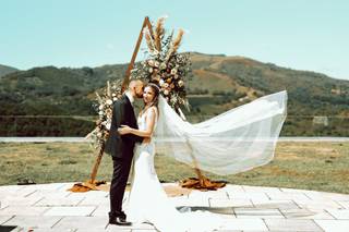 Pareja vestida de novios delante de una estructura de madera triangular con muchas flores