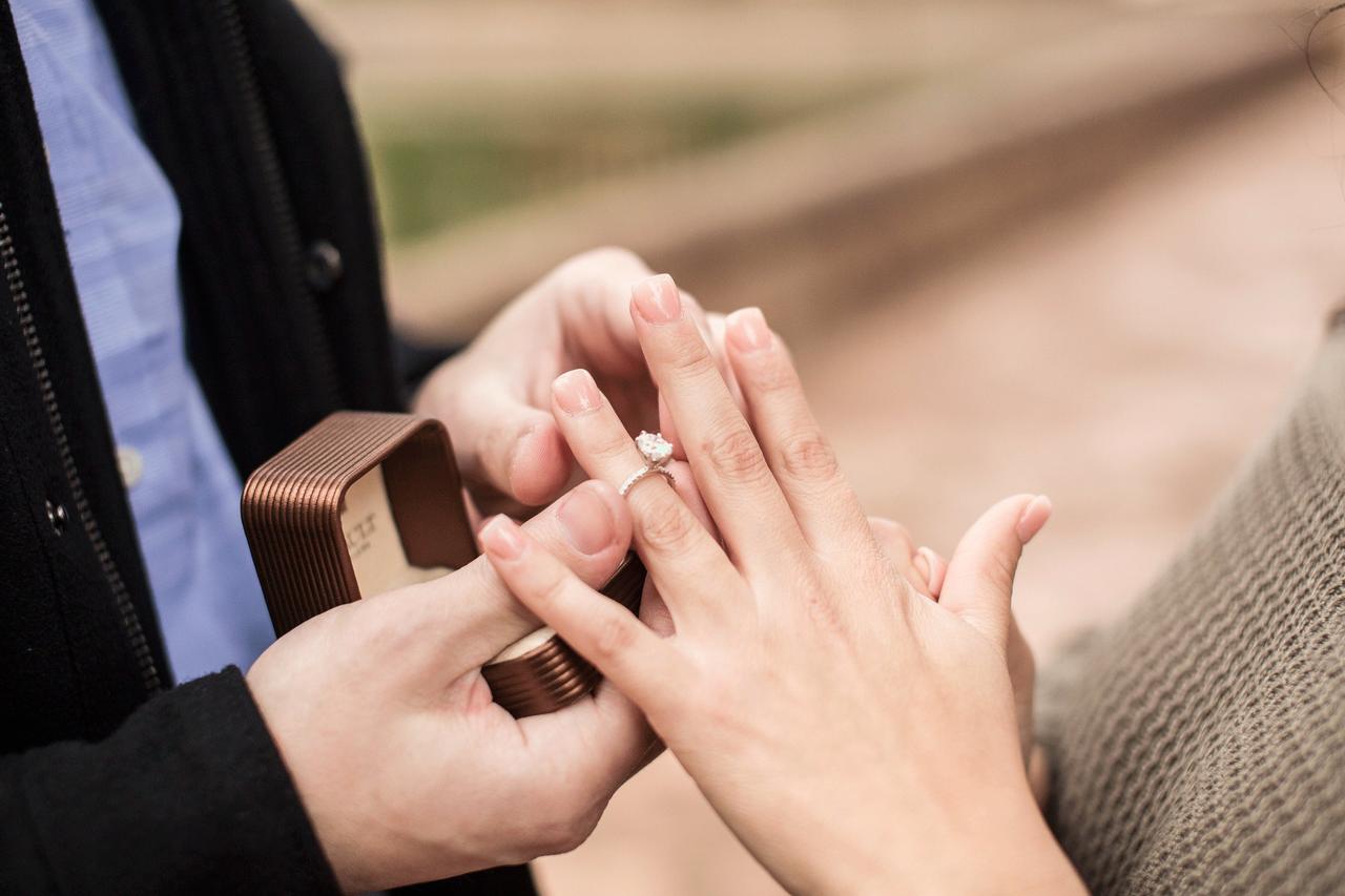 Manos de hombre sujetando una cajita mientras le ponen un precioso anillo con diamantes en el dedo anular de la mano izquierda