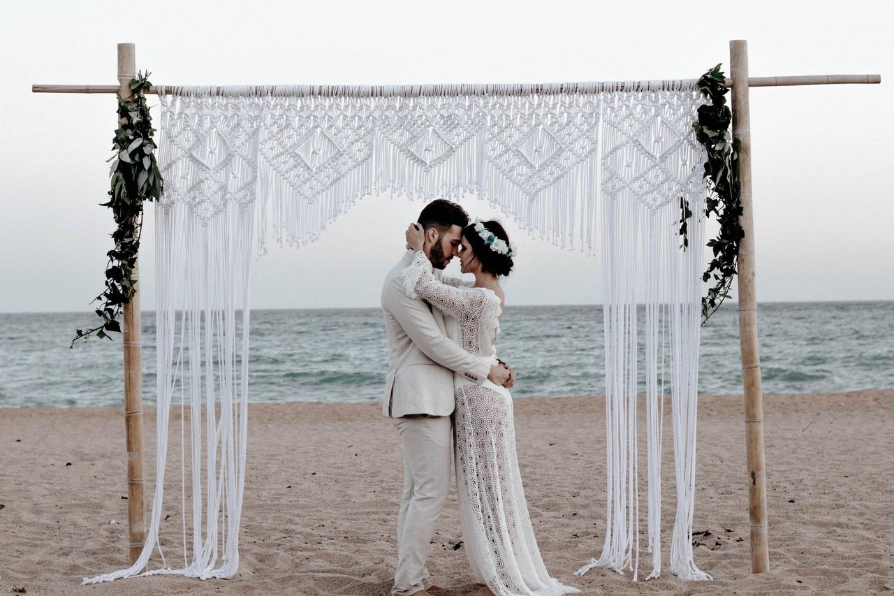 Pareja besándose antes su altar de boda en la playa hecho con madera, flores y macramé