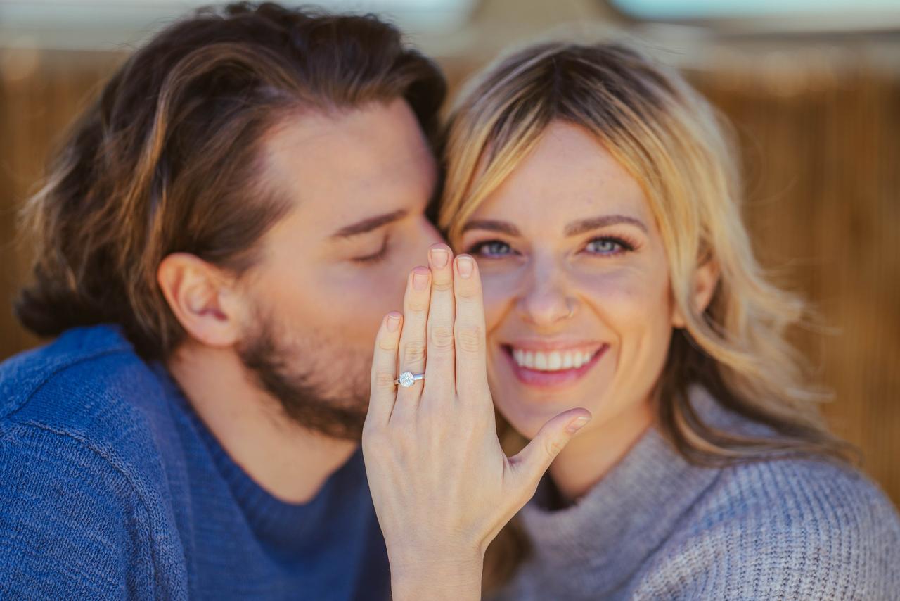 Chico le da un beso a una chica rubia y muy sonriente que enseña a cámara su mano con un precioso anillo en el dedo anular de la mano izquierda