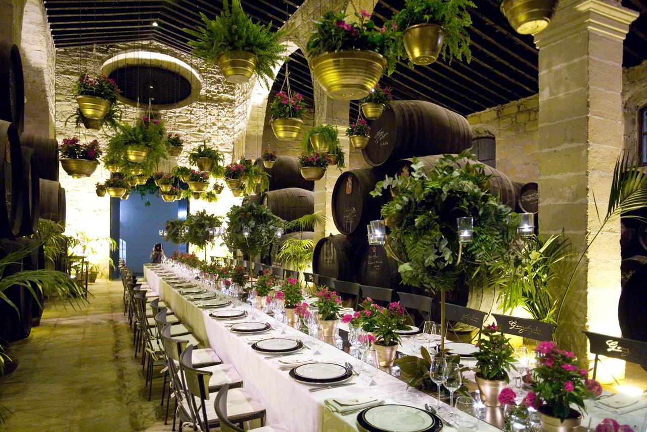 Interior de una bodega con barriles de vino y una larga mesa horizontal preparada para una comida o cena elegante