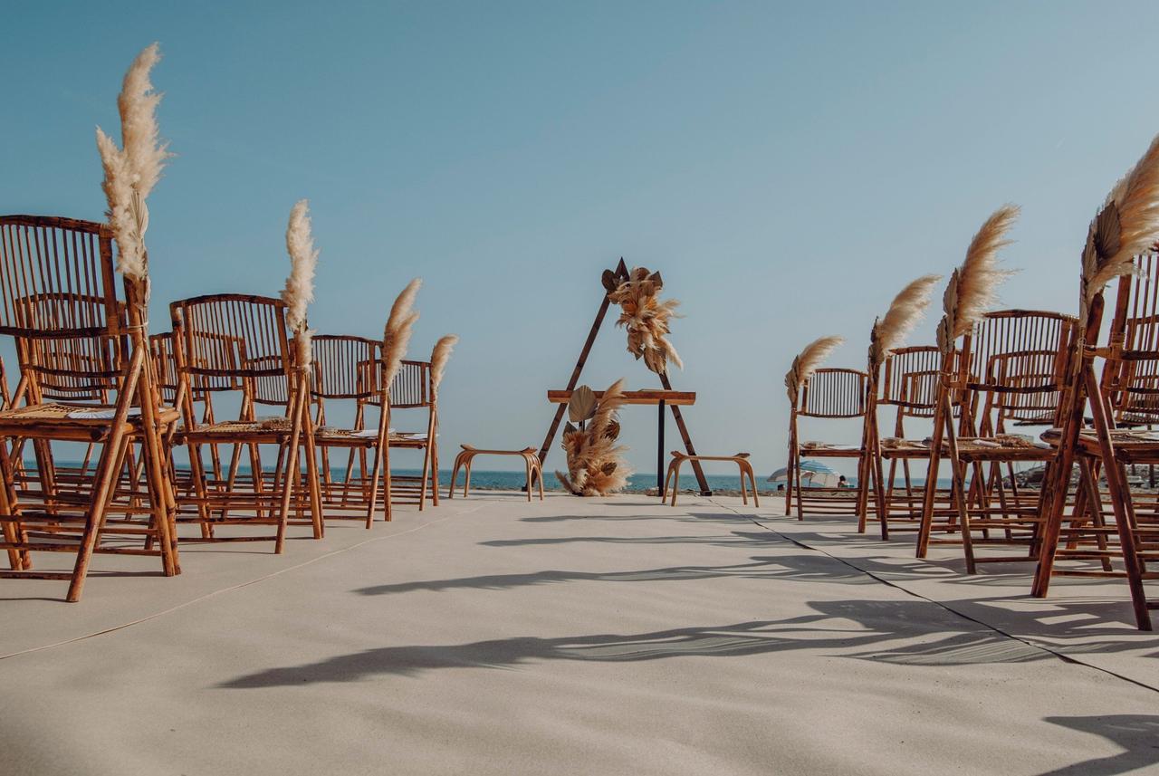 Decoración para bodas en la playa con sillas plegables de madera, dos banquetas y un altar con forma de triángulo