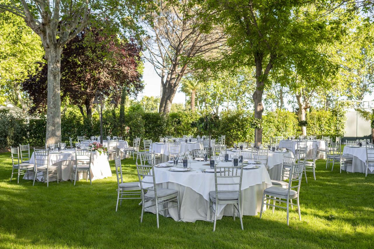 Jardín con césped y muchos árboles lleno de mesas redondas preparadas para una comida o cena nupcial