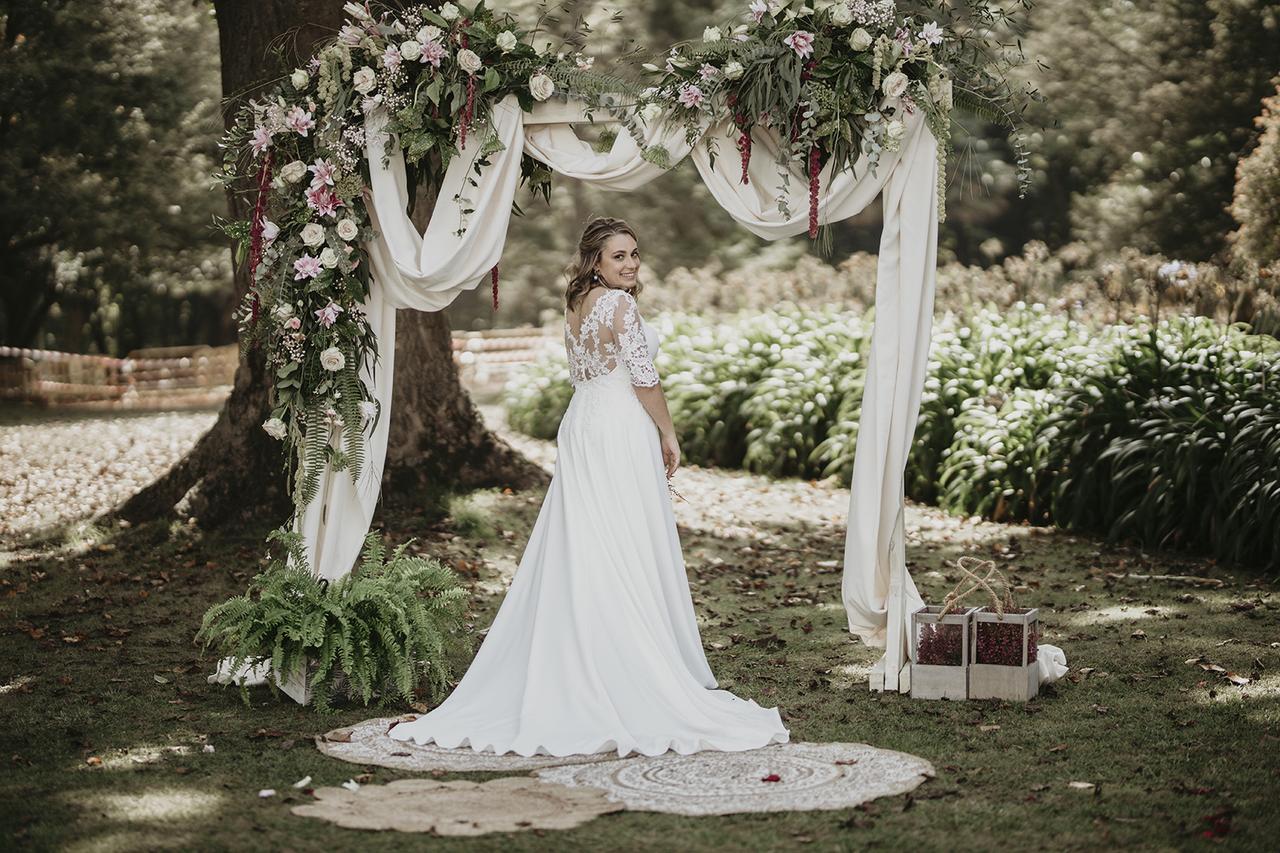 Chica vestida de novia de lado delante de un arco con telas y variados tipos de flores