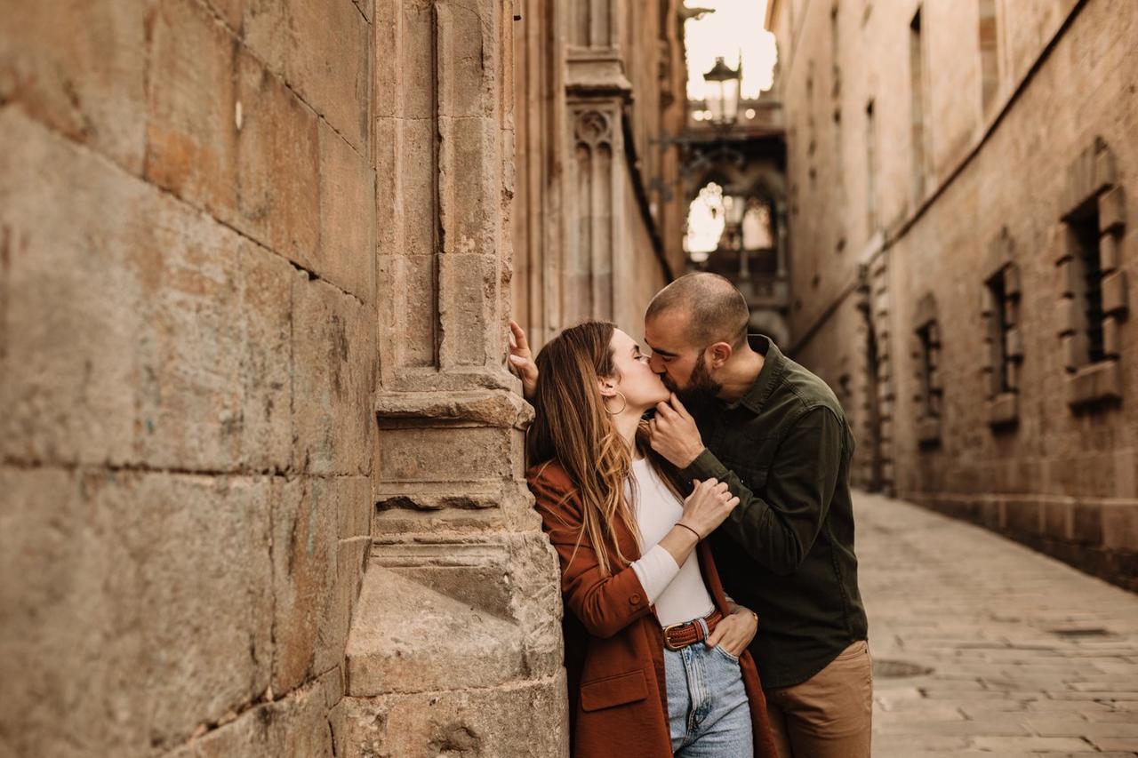Chico con barba y chica con la melena suelta se besan en una calle, apoyados en las paredes de uno de los edificios