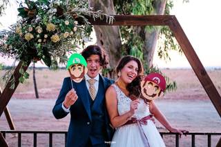 Chico con traje y chica vestida de novia sujetan en sus manos unas caretas de Mario bros debajo de una estructura de madera hexagonal al aire libre
