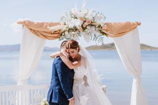 Pareja abrazada en el altar de su boda civil al aire libre delante de una arco con telas de color blanco y muchas flores en el centro