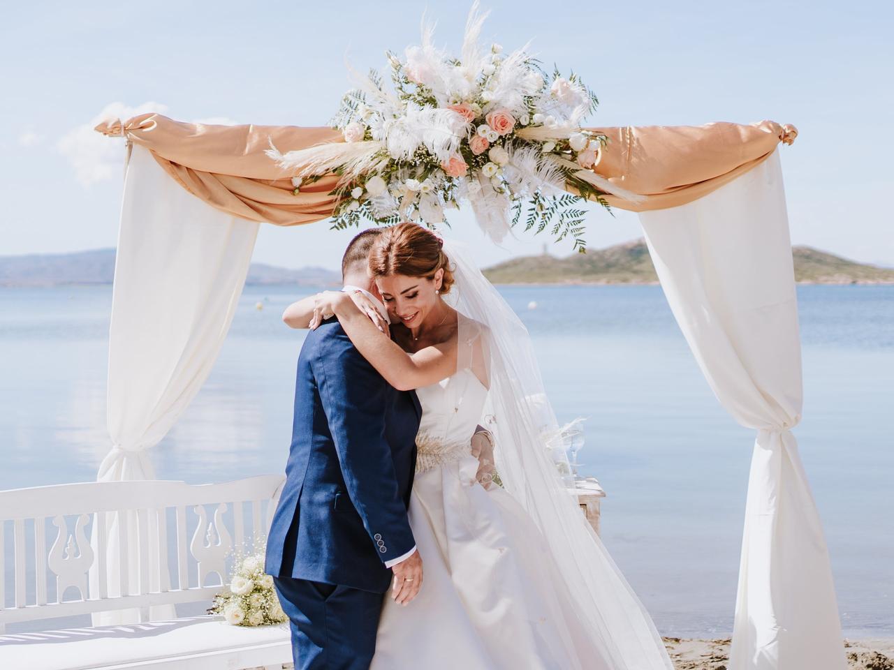 Pareja abrazada en el altar de su boda civil al aire libre delante de una arco con telas de color blanco y muchas flores en el centro