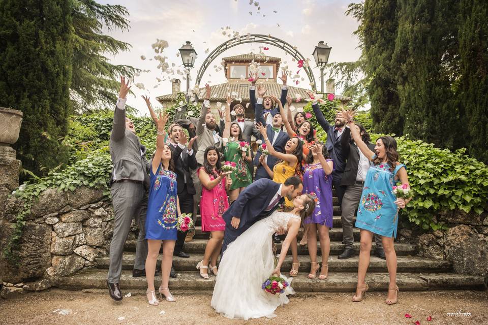 Grupo de invitados muy sonrientes en una escalera tiran pétalos de flores al aire mientras una pareja de recién casados se besa