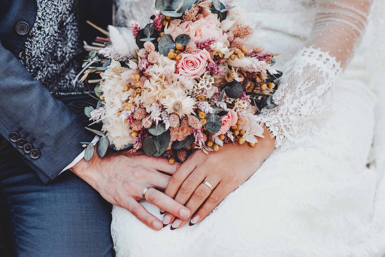 Qué se siente estar enamorado: manos entrelazadas de hombre con traje y de mujer con vestido de novia, con un bonito ramo de flores entre ellas